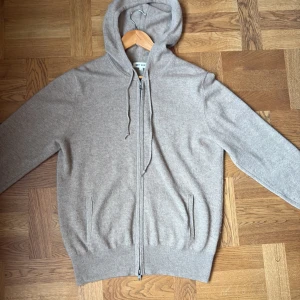 Beige hoodie i kashmir från Soft Goat - Mjuk och lyxig beige hoodie från Soft Goat i kashmir. Tröjan har huva med snörning, hel dragkedja framtill och ribbade muddar. Perfekt för dig som gillar chill och stilren look. Långärmad och med två fickor på sidorna. Färgen är hazelnut / beige. Den är i nyskick men använd. Tredje bilden ser man att tyget har öppnat men syns inte vid användning  och påverkar inte dragkedjan det minsta. Vid fler frågor är det bara att höra av sig!