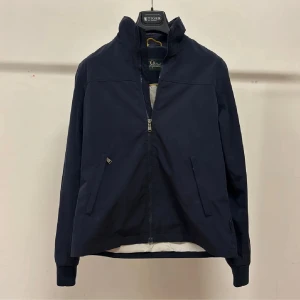 Mörkblå vindjacka Peak Performance M - Snygg mörkblå vindjacka från Peak Performance med ståkrage och dragkedja framtill. Jackan har två snedställda fickor, ribbade muddar och detaljer i vitt och gult på insidan. Perfekt för dig som gillar stilren och sportig look.