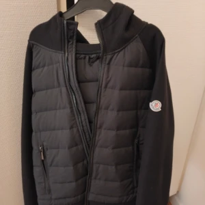 Svart cardigan  från Moncler - Svart pufferjacka från Moncler med quiltad framsida och huva. Jackan har dragkedja, två sidofickor med dragkedja och Moncler-logga på ärmen. Materialet är en mix av polyester och stickad tyg på ärmar och axlar. Perfekt för kyliga dagar.