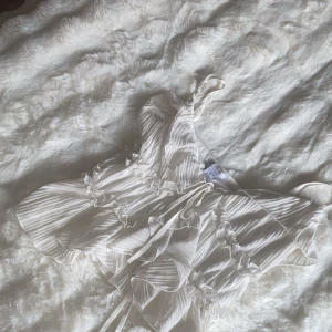 Vit omlott blus med volanger - Säljer en superfin vit omlott blus med tunna axelband, volanger och knyt framtill. Blusen har ett randigt mönster och är gjord i ett lätt, transparent syntetmaterial som ger en luftig känsla. Perfekt för dig som gillar romantiska detaljer och flowy vibes.