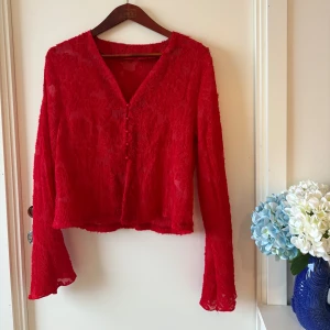 Röd blus med pärlknappar och v-ringning - Röd romantisk blus med v-ringning och dekorativa pärlknappar framtill. Blusen har lång ärm och är gjord i ett lätt transparent, strukturerat tyg med blommiga detaljer. Croppad passform som ger en cool vibe. Perfekt för dig som vill sticka ut.