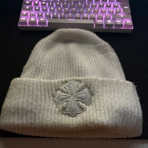 Vit ribbad mössa med korsdetalj - Säljer en vit ribbad mössa från Chrome Hearts med broderad korsdetalj framtill och en metallblomma på toppen. Mössan är stickad och har uppvikt kant för extra stilpoäng. Perfekt för dig som gillar streetwear med exklusiva detaljer.