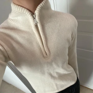 Beige/vit stickad half zip från Peak Performance - Stickad tröja från Peak Performance med half zip och hög krage. Tröjan är i mjuk ullblandning, har lång ärm och en clean, klassisk look. Perfekt för lager-på-lager och chillare dagar.