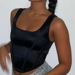 Svart glansig korsett-topp -  svart korsett-topp med glansigt tyg och breda axelband. Toppen har en figurnära passform och markerade sömmar framtill