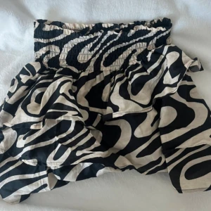 Svartvit zebramönstrad kortkjol - Supersnygg kortkjol med svartvitt zebramönster och bred smock i midjan. Kjolen har volanger som ger extra volym och en cool look. Perfekt för dig som vill sticka ut med ett djurmönster och gillar trendiga detaljer.