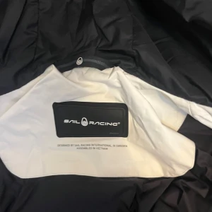 Vit pufferjacka från Sail Racing - Säljer en vit pufferjacka från Sail Racing med svarta detaljer och huva. Jackan har dragkedja framtill, logga på bröstet och en patch på ärmen. Perfekt för kalla dagar och har en sportig look.