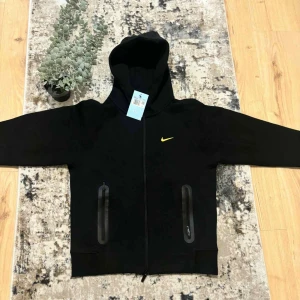 Svart NOCTA Nike hoodie med dragkedja - Svart hoodie från Nike NOCTA med gul swoosh på bröstet och broderade detaljer bak. Tröjan har hel dragkedja, två fickor med dragkedja och huva. Materialet är mjukt och känns premium, perfekt för en clean och sportig look.