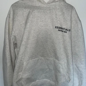 Grå Essentials Fear of God hoodie - Säljer en stilren ljusgrå hoodie från Essentials Fear of God. Tröjan har huva, magficka och svart logga tryckt på bröstet. Perfekt för dig som gillar streetwear och vill ha en enkel men snygg look.