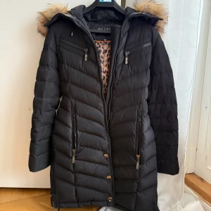 Svart dunjacka med päls från Hollies - Snygg svart dunjacka från Hollies med fluffig päls på huvan och coola detaljer som dragkedjor och bälte i midjan. Jackan har quiltad design, långa ärmar och leopardmönstrat foder. Perfekt för kalla dagar när du vill vara både varm och stilren. Jackan har små defekter, den har sminkfläckar vid kragen som jag tyvärr inte tror går bord. Den har även en reva vid ena knäppet nere på sidan liksom, inget som stör funktionen men den finns där. Annars fin!