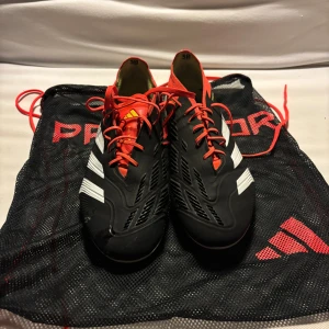 Adidas Predator fotbollsskor svart/röd - Säljer ett par Adidas Predator fotbollsskor i svart med röda och vita detaljer. Skorna har snörning, mönstrad ovandel och dobbar för gräsplan. Kommer med original Predator-påse. Perfekta för dig som vill sticka ut på planen.