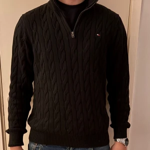 Svart stickad half zip från Tommy Hilfiger - Svart stickad tröja från Tommy Hilfiger med half zip och kabelstickat mönster. Liten logga på bröstet och ribbade muddar vid ärmslut och nederkant. Hög krage med Hilfigersignatur på insidan. Perfekt för dig som gillar klassisk stil.
