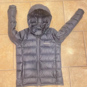 Mörkblå dunjacka Peak Performance - Snygg mörkblå dunjacka från Peak Performance med huva och dragkedja framtill. Jackan har ett quiltat mönster och är perfekt för kalla dagar. Långärmad modell med stilren logga på bröstet. Passar dig som vill ha en varm och cool jacka.