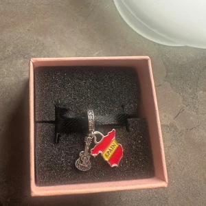 Charm till armband med Spanien-tema - Unik charm till armband i silverfärgad metall med två hängen: en gitarr och en röd-gul Spanien-karta med texten 'SPAIN' på framsidan och 'I love Spain' på baksidan. Perfekt för dig som älskar musik och resor! Säljs i fin presentask.