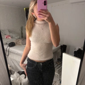 Ljusbeige stickad ärmlös polotopp - Supermysig ljusbeige stickad polotopp med ärmlös design och mjuk, fluffig känsla. Toppen har en hög hals och sitter snyggt figurnära. Perfekt att styla med jeans eller kjol för en trendig look.