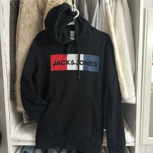 Svart hoodie från Jack & Jones - Svart hoodie från Jack & Jones med stor logga framtill i rött, vitt och blått. Tröjan har huva med snörning och en klassisk känguruficka. Tillverkad i mjuk bomullsmix, perfekt för chill dagar eller när du vill ha en clean streetstyle.