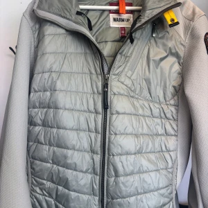 Parajumpers Warm Up Jacket – grå lätt jacka herr M - Superfin lättviktsjacka från Parajumpers i modellen Warm Up. Färg: ljusgrå/silvergrå. Perfekt till vår, höst eller som mellanlager under en skaljacka.  Jackan har quiltad framsida med lätt vaddering, stretchiga ärmar och sidor för bästa komfort. Klassisk PJS-logga på ärmen och gul remdetalj vid kragen.