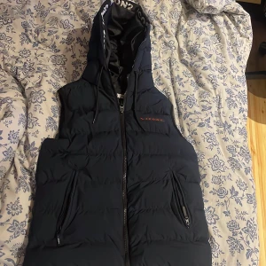 Svart dunväst med huva från ICON2 - Svart dunväst från ICON2 med stor huva och dragkedja framtill. Huva med tryckt logga längs kanten och två fickor med dragkedja. Västen har en quiltad design och en liten röd logga på bröstet. Perfekt för lager-på-lager och streetstyle.