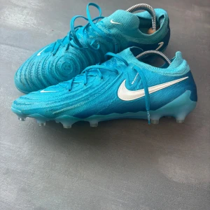 NIKE PHANTOM GX II ELITE AG MAD AMBITION - BLÅ/VIT - Nike Phantom GX Elite DF AG – toppmodell i Nikes Phantom-serie, använd av flera proffsspelare. Gripknit-överdel för perfekt bollkänsla och Dynamic Fit-krage för stöd runt ankeln. Använda men i gott skick, rengjorda. Storlek 44 EU