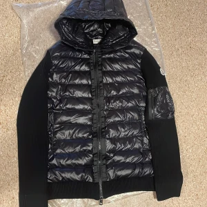 Svart pufferjacka från Moncler - Svart pufferjacka cardigan från Moncler med glansig quiltad front och huva, stickade ärmar och rygg. Jackan har dragkedja framtill och en ficka med dragkedja på ärmen. Perfekt för dig som gillar stilren design och exklusiva detaljer.