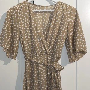 Beige omlottklänning med vita prickar - Superfin beige omlottklänning med vita prickar och knyt i midjan. Klänningen har v-ringning, korta vida ärmar och ett luftigt fall. Perfekt för varma dagar och ger en avslappnad men ändå trendig look.