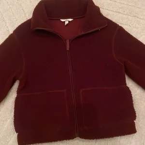 Mörkröd teddyjacka från H&M - Säljer en mörkröd teddyjacka från H&M i storlek S. Jackan har en hög krage, två stora fickor framtill och stängs med dragkedja. Materialet är mjukt och fluffigt, perfekt för kyliga dagar. Enkel och stilren design som är lätt att matcha. Den är aldrig andvänd, bara testad men prislappen finns inte kvar.