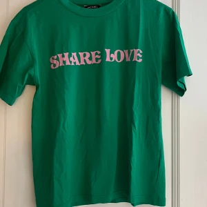 Grön t-shirt Monki Share Love - Grön t-shirt från Monki med trycket 'SHARE LOVE' i rosa bokstäver på bröstet. Klassisk passform med rund hals. 