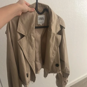 Croppad beige trenchcoat från Zara - Säljer en croppad trenchcoat från Zara i beige med breda slag, svarta knappar och coola detaljer vid ärmarna. Jackan har en loose passform och är perfekt för dig som gillar trendiga och unika plagg. Materialet är bomull och syntet.