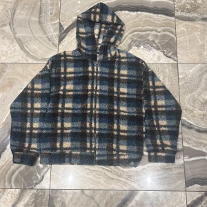 Rutig fleecehoodie från Lindex - Mysig hoodie från Lindex i rutig design med blå, beige och svarta toner. Tillverkad i mjuk fleece med dragkedja framtill och huva. Perfekt för kyliga dagar när du vill vara både varm och stilren.kontakta mongol du vill köpa