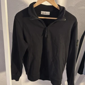 Svart halvzip tröja från Stone Island - Säljer en svart långärmad tröja från Stone Island med halv dragkedja och klassisk logotyp på ärmen. Tröjan har en normal passform och är tillverkad i mjukt bomullsmaterial. Perfekt för en stilren och avslappnad look. Väldigt liten defekt på sista bilden men är ihopsydd och påverkar inte något.