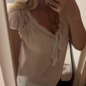Vit blus med spets och knyt - Säljer en söt vit blus med korta ärmar och spetsdetaljer vid axlarna. Blusen har en v-ringad hals med knyt framtill och är gjord i ett lätt, luftigt material! Skriv med fler frågor eller bilder💗