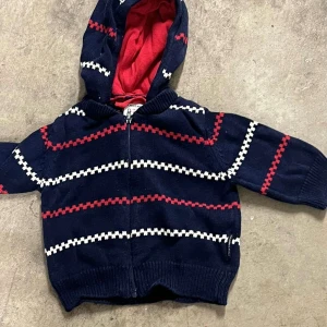 Marinblå hoodie med ränder och huva - Säljer en marinblå hoodie från KappAhl med röda och vita randiga detaljer. Tröjan har dragkedja framtill, ribbade muddar och en mysig huva med röd insida. Perfekt för kyliga dagar och enkel att slänga på sig.