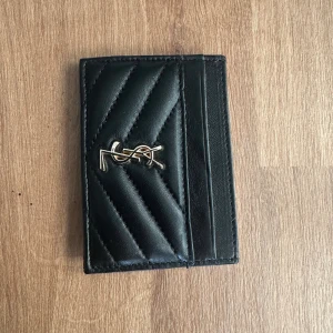 Svart quiltad plånbok från YSL - Snygg svart plånbok från YSL i skinn med quiltad design och guldigt YSL-emblem på framsidan. Plånboken har flera kortfack och stilrena diagonala sömmar som ger en lyxig känsla. Perfekt för dig som gillar klassisk och trendig stil.