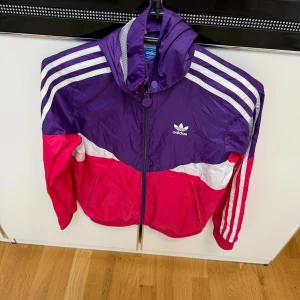 Adidas färgglad vindjacka med huva - Säljer en snygg Adidas vindjacka i lila, rosa och vitt med klassiska ränder på ärmarna. Jackan har huva, dragkedja framtill och två fickor med zip. Perfekt för dig som gillar sportig stil och färg. Adidas-logga på bröstet och meshfoder i huvan.
