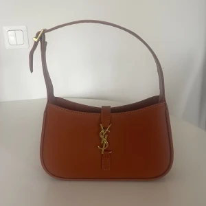 Brun axelväska från Yves Saint Laurent - Snygg brun axelväska från Yves Saint Laurent i slätt skinn med guldigt YSL-logo framtill. Väskan har justerbart axelband och stilren, avlång form. 
