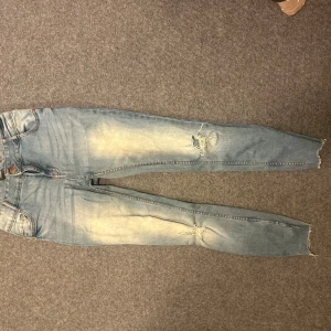 Ljusblå skinny jeans med slitningar - Säljer ett par ljusblå skinny jeans med slitningar på knäna och råa benslut. Jeansen har klassisk femficksdesign och är tillverkade i stretchig denim för en tight passform. Perfekta för en avslappnad och trendig look.