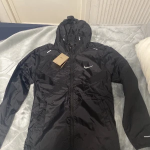 Svart Nike vindjacka med huva M - Svart vindjacka från Nike i storlek M, med huva och hel dragkedja framtill. Jackan har vita Nike-loggor på bröstet och axlarna, samt praktiska fickor på sidorna. Materialet är lätt och syntetiskt, perfekt för blåsiga dagar. Snygg och sportig look.