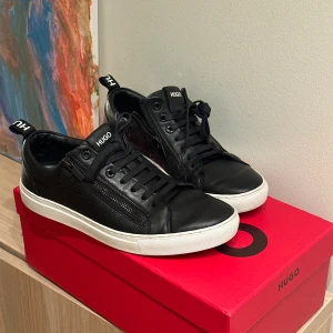 Svarta sneakers från Hugo Boss storlek 41 - Snygga svarta sneakers från Hugo Boss med vit sula, perforerade sidopaneler och dragkedjedetalj. Skorna har snörning, rund tå och är tillverkade i skinn. Diskret Hugo-logga på plös och hälflik. Perfekta för en clean och stilren look. Storlek 41 modell: Futurism tenn