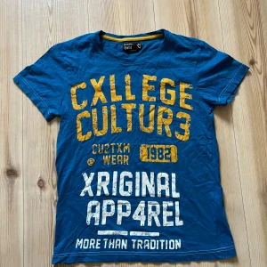 Blå t-shirt  från Solid strl M - Blå t-shirt från Solid i storlek medium med stort college-inspirerat tryck i gult och vitt på framsidan. Rund hals med gul kantdetalj. Skön bomullskvalitet och korta ärmar. Perfekt för dig som gillar streetstyle och tydliga prints.