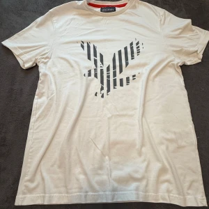 Vit t-shirt med randigt tryck Lyle & Scott - Vit t-shirt från Lyle & Scott i storlek M. Framsidan har ett stort svart tryck med randigt mönster i form av en fågel. Klassisk rund hals med röd insida. Mjuk och skön bomullskvalitet, perfekt till jeans eller shorts.