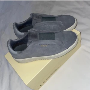 Grå sneakers från Axel Arigato - Snygga grå sneakers från Axel Arigato i mocka med vit platt sula och elastiskt band över vristen. Minimalistisk design med guldfärgad logga på sidan. Perfekta för dig som gillar stilrena och moderna skor.