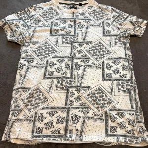 Vit t-shirt med paisley från Brave Soul - Säljer en vit t-shirt från Brave Soul med svart paisley- och rutmönster över hela plagget. T-shirten har korta ärmar och är gjord i mjuk bomull. Snyggt tryck med detaljerade rutor och prickar som ger en unik vibe.