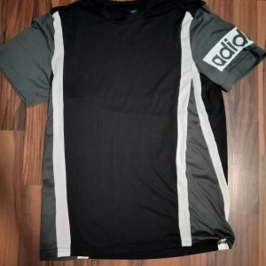 Svart/grå Adidas t-shirt Climalite S - Snygg t-shirt från Adidas i svart och grått med vita vertikala ränder längs sidorna. Kortärmad modell med stort Adidas-tryck på ena ärmen. Tillverkad i lätt och funktionell polyester Climalite som andas, perfekt för träning eller chill.