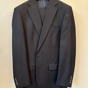 Mörkblå kostym Dressmann Stl 46 W31/L32 - Klassisk svart kostym från Dressmann med kavaj och matchande byxor. Kavajen har snygga slag, två framfickor med lock och fyra knappar vid ärmslutet. Kostymen är tillverkad i en blandning av polyester och viskos för en stilren look och skön känsla. Kavajen har Stl 46 och byxorna W31/L32. Byxorna har lagts upp en bit för att passa mig som är 178cm lång. Väldigt sparsamt använd. 