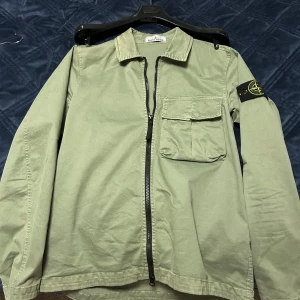 Grön Stone Island overshirt - Snygg ljusgrön overshirt från Stone Island med klassisk krage, bröstficka och svart logotyp-patch på ärmen. Använt 1-2 gånger. Stone Island galge tillkommer även. Priset kan diskuteras.