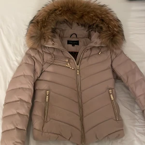 Beige pufferjacka med fuskpäls från Hollies - Snygg beige pufferjacka från Hollies med stor huva och fluffig fuskpäls. Jackan har guldfärgade dragkedjor, flera fickor och ett emblem på ärmen. Perfekt för kalla dagar och riktigt trendig look.