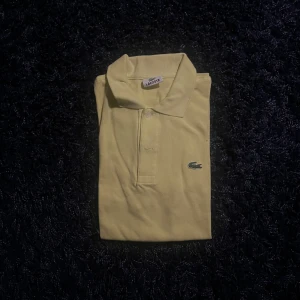 Gul pikétröja från Lacoste - Snygg ljusgul pikétröja från Lacoste med klassisk krage och två knappar framtill. Ikoniska krokodilloggan broderad på bröstet. Perfekt för dig som vill ha en fräsch och stilren look. Tillverkad i mjuk bomull som andas.