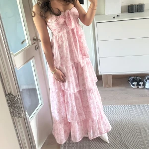 Balklänning!! Ljusrosa blommig långklänning med volanger -  ljusrosa långklänning med blommigt mönster och flera lager volanger. Axelbandslös modell med söt rosett framtill och flowig passform. Perfekt för sommarens alla tillfällen när du vill känna dig extra fin. Har sytt upp den då den var för lång, skulle säga att den passar även kortare personer. 155-170. Pris går att diskuteras.