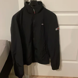 Svart vindjacka från Tommy Jeans - Svart vindjacka från Tommy Jeans med klassisk logga på bröstet och ärmen. Jackan har hög krage, dragkedja framtill och är tillverkad i ett lätt syntetmaterial. Perfekt för dig som gillar stilrena och sportiga plagg med diskreta detaljer.