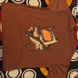 Brun Domo t-shirt med coolt tryck - Säljer en brun t-shirt från Domo med ett stort, grafiskt tryck av Domo-figuren som spelar elgitarr i orange och beige toner. T-shirten har korta ärmar och är gjord i mjuk bomull, perfekt för dig som gillar unika motiv och streetstyle.