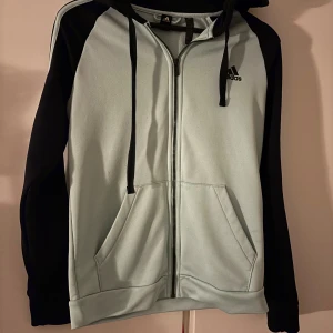 Mörkblå och ljusblå hoodie från Adidas - Snygg hoodie från Adidas i ljusblått med mörkblå ärmar och huva. Tröjan har dragkedja, snörning vid huvan och två stora fickor fram. Adidas-logga på bröstet och klassiska ränder på axlarna. Perfekt för chill eller träning.